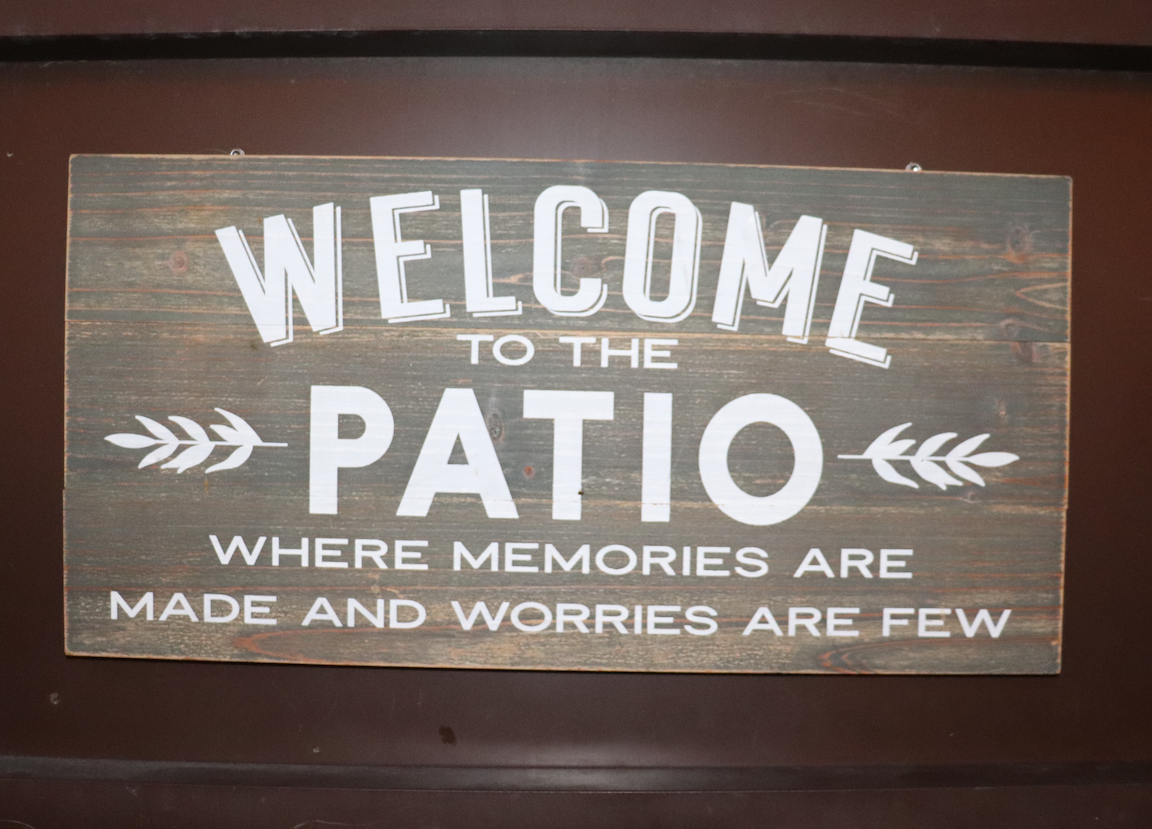 Dark wooden sign with white letters saying "Welcome to the Patio - where memories are made and worries are few"